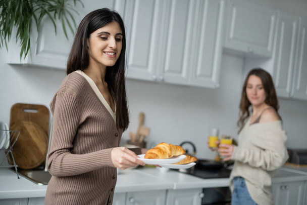 Lesbian woman holding croissants near blurred girlfriend in kitchen  - Foto, Imagem