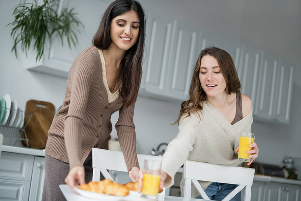 Happy same sex couple putting orange juice and croissants on table in kitchen  - Fotó, kép