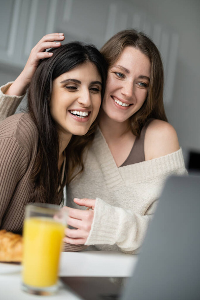 Sonriente pareja de lesbianas mirando a la computadora portátil cerca borrosa desayuno en la cocina  - Foto, Imagen