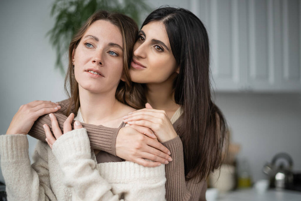 Lesbian women in warm clothes hugging and looking away in kitchen  - Foto, Bild
