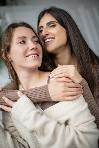 Cheerful lesbian woman embracing partner in warm sweater at home  - Fotoğraf, Görsel