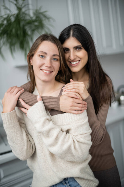 Positive same sex couple in warm clothes hugging in kitchen  - Φωτογραφία, εικόνα