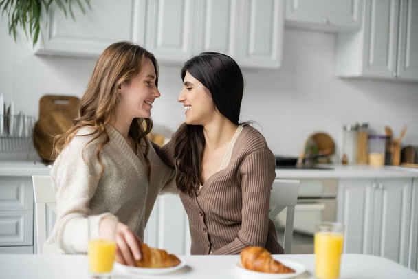 Vista lateral de pareja lesbiana positiva mirándose el uno al otro cerca del desayuno en la cocina  - Foto, Imagen