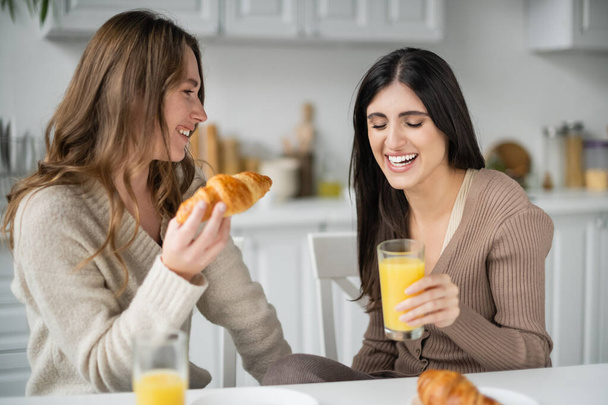 Positive same sex couple holding croissant and orange juice during breakfast in kitchen  - Foto, Bild
