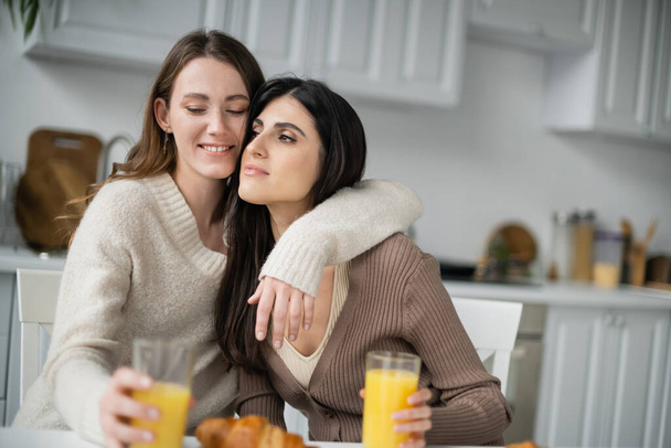 Young lesbian woman hugging girlfriend near orange juice and croissant in kitchen  - Фото, изображение