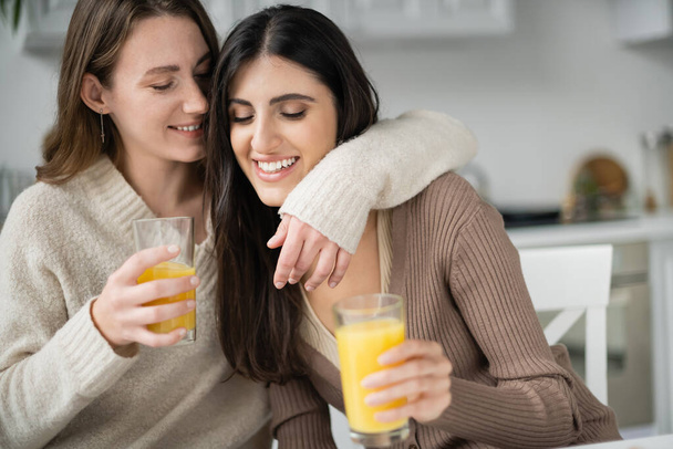Young woman in sweater hugging girlfriend and holding orange juice in kitchen  - Φωτογραφία, εικόνα