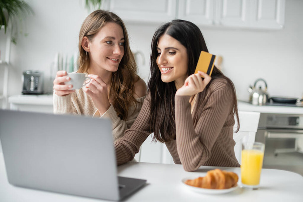 Pareja lesbiana usando laptop y tarjeta de crédito durante el desayuno en la cocina  - Foto, Imagen