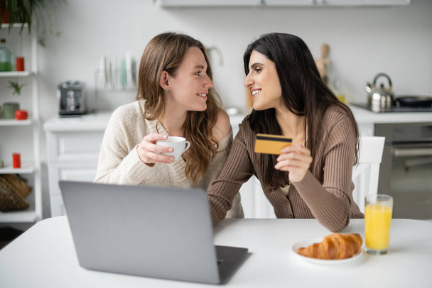 Widok z boku uśmiechnięta para tej samej płci posiadający kartę kredytową i kawę w pobliżu laptopa i śniadanie w kuchni  - Zdjęcie, obraz