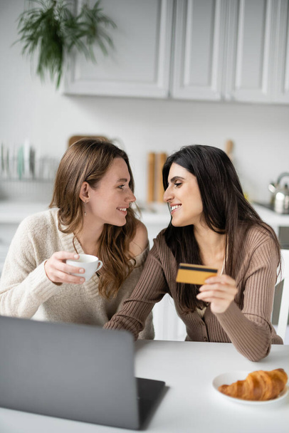 Widok z boku pozytywnej pary lesbijek trzymających kawę i kartę kredytową w pobliżu zamazanego laptopa i rogalika w kuchni  - Zdjęcie, obraz