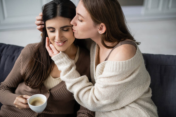 Lesbian woman in warm sweater kissing girlfriend with cup of coffee on couch  - Fotó, kép