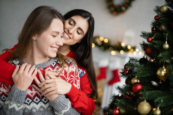 glimlachende brunette vrouw knuffelen vriendin tijdens kerst feest thuis  - Foto, afbeelding