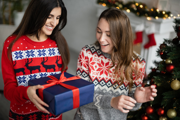 Femme souriante donnant cadeau à petite amie près de l'arbre de Noël à la maison  - Photo, image