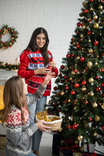 Positive woman holding christmas ball near girlfriend and pine tree in living room  - Photo, Image