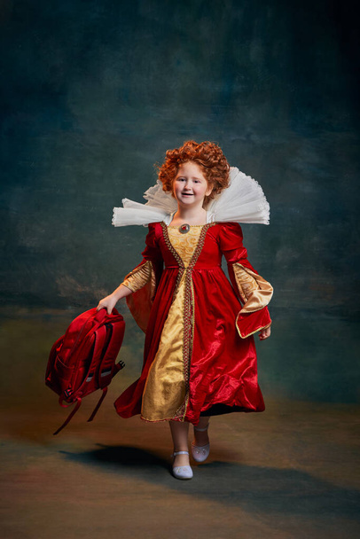 Portrait of little red-headed girl, child in costume of royal person isolated on dark green background. School time. Concept of historical remake, comparison of eras, medieval fashion, emotions, queen - Photo, image