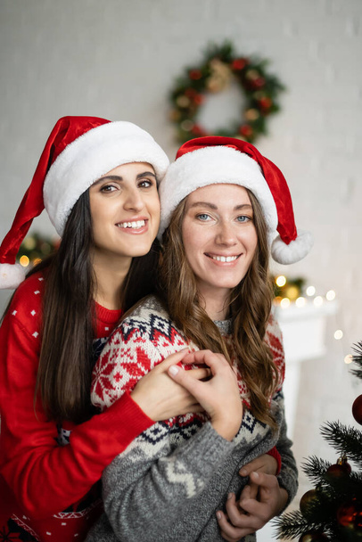 Happy lesbian couple in santa hats and sweaters holding hands at home  - Photo, Image