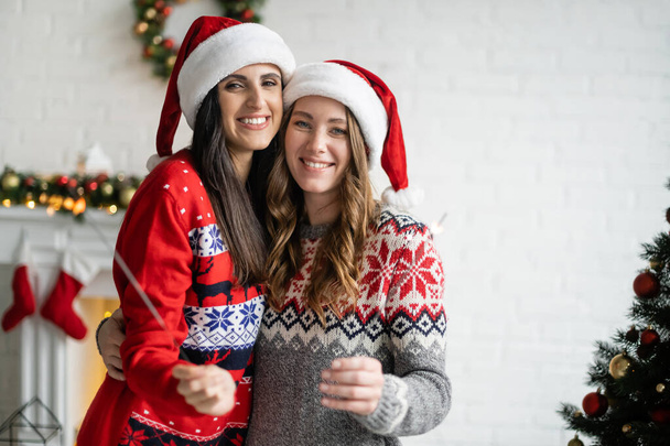 Glückliches gleichgeschlechtliches Paar mit Weihnachtsmann-Hüten, Wunderkerzen in der Hand und Blick in die Kamera zu Hause  - Foto, Bild