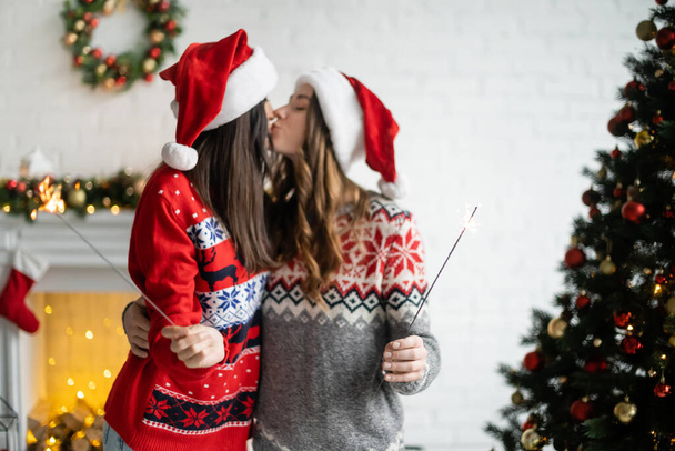 Side view of blurred lesbian couple in santa hats kissing and holding sparklers at home  - Фото, изображение