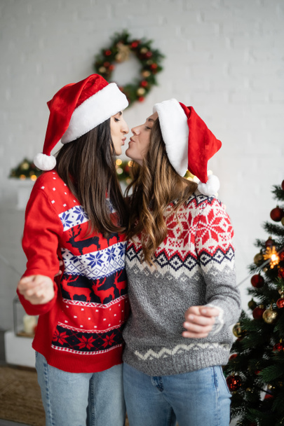 Side view of same sex couple in santa hats kissing and holding sparklers in living room  - Фото, зображення