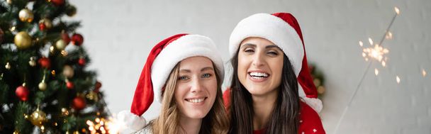 Cheerful lesbian couple in santa hats looking at camera near sparklers and christmas tree at home, banner  - Photo, Image