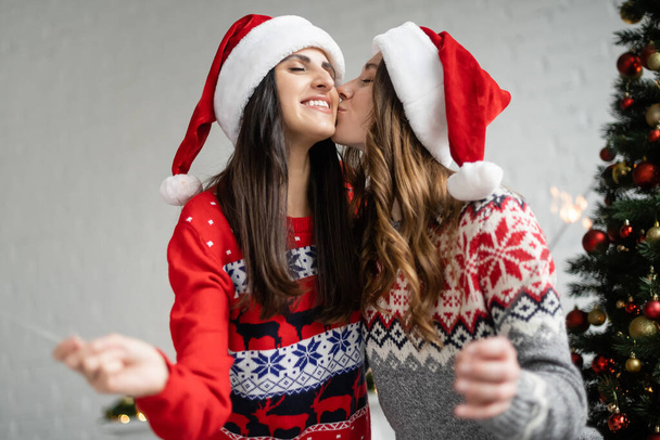 Young lesbian woman in sweater and santa hat kissing girlfriend with sparkler at home  - Photo, Image