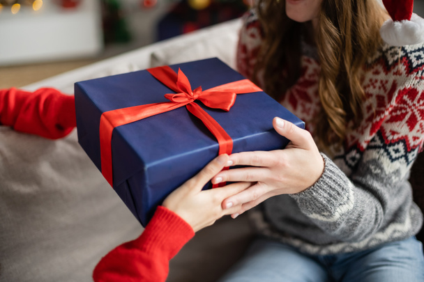 Cropped view of woman in sweater giving christmas present to girlfriend on couch at home  - Photo, Image