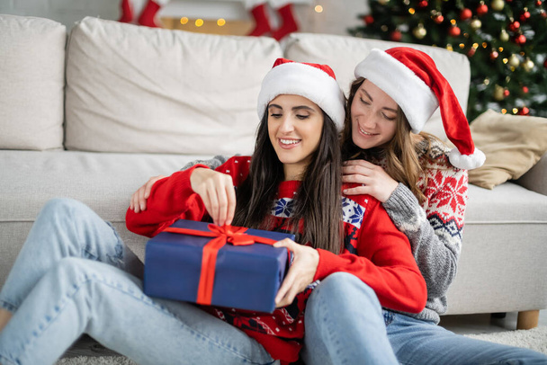 Smiling woman in santa hat hugging girlfriend with christmas gift in living room  - Zdjęcie, obraz