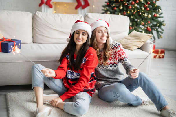 Junges lesbisches Paar mit Weihnachtsmann-Hüten und Wunderkerzen zu Hause  - Foto, Bild