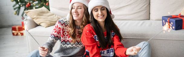 Giovane coppia dello stesso sesso in cappelli di Babbo Natale che tengono scintille in soggiorno, banner  - Foto, immagini