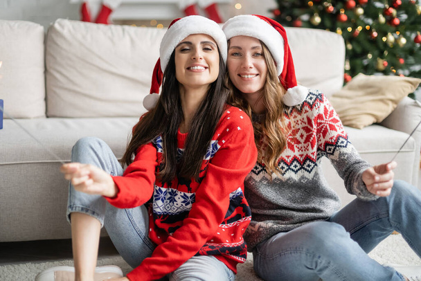 Positive lesbian couple in santa hats holding sparklers in living room  - Foto, Bild