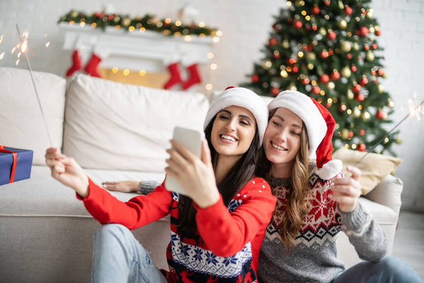 Cheerful same sex couple in santa hats taking selfie on smartphone and holding sparklers at home  - Photo, image