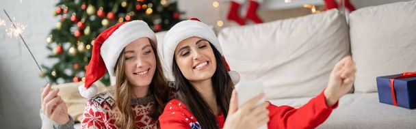 Χαρούμενο ζευγάρι λεσβιών με καπέλα και πουλόβερ Σάντα χρησιμοποιώντας smartphone και κρατώντας sparklers στο σπίτι, πανό  - Φωτογραφία, εικόνα