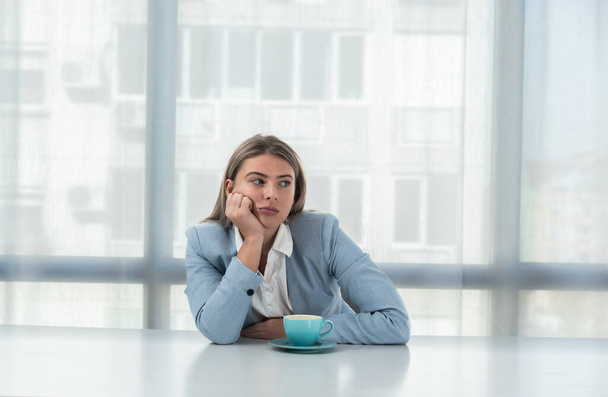 But first coffee. Young tired business woman in formal wear sitting in the office at empty desk with cup of coffee. Female white collar worker, freelancer financial expert morning routine. - Photo, Image