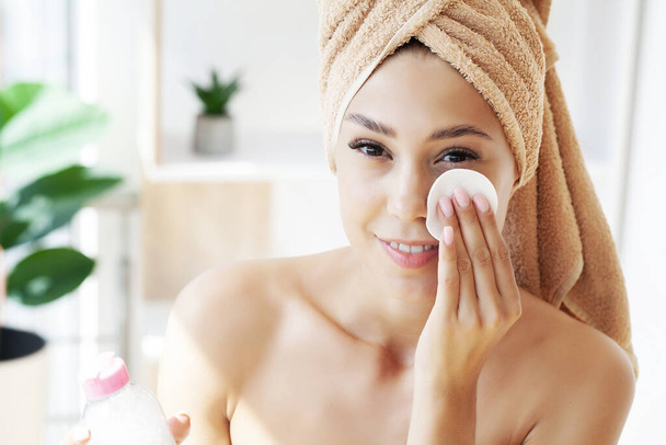 Hermosa mujer está limpiando su cara usando un disco de algodón mientras se mira en el espejo en el baño. - Foto, imagen