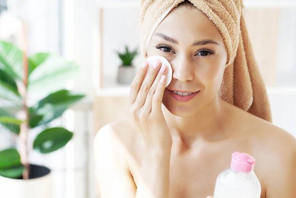 Όμορφη γυναίκα καθαρίζει το πρόσωπό της χρησιμοποιώντας ένα δίσκο βαμβάκι, ενώ κοιτάζοντας στον καθρέφτη στο μπάνιο. - Φωτογραφία, εικόνα
