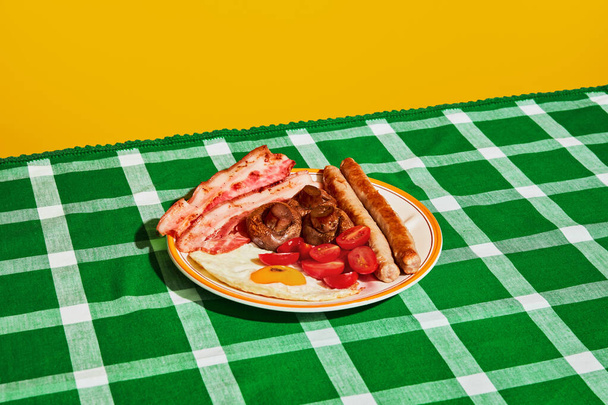Plate of delicious English breakfast with fried eggs, bacon, sausage, mushrooms and tomato on green tablecloth. Vintage, retro style. Food pop art. Complementary colors. Copy space for ad, text - Photo, Image