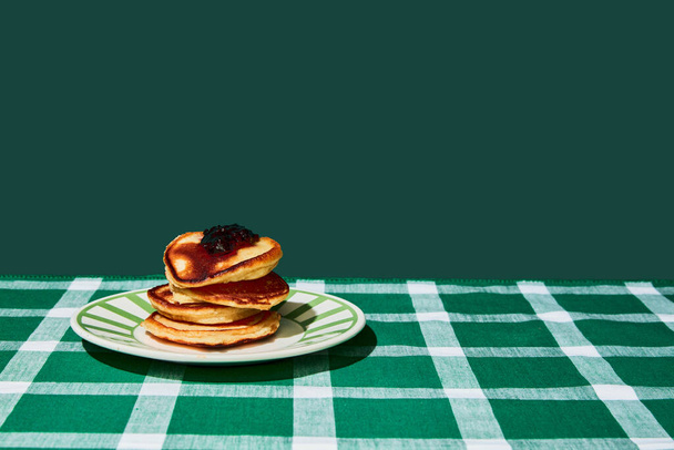 Plate of delicious sweet pancakes with jam on green tablecloth over green background. Vintage, retro style interior. Food pop art photography. Complementary colors. Copy space for ad, text - Photo, image