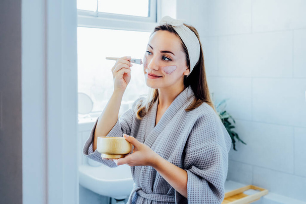 Young woman in bathrobe looking in the mirror and applying facial natural cosmetic clay mask on her face in bathroom. Cosmetic procedures for skin care at home. Beauty self-care. Selective focus. - Foto, afbeelding