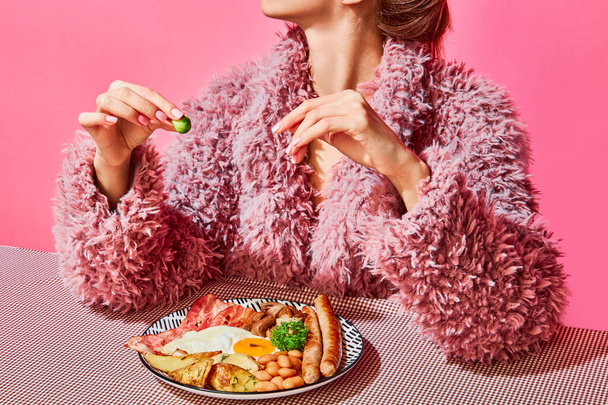 Woman in furry pink coat eating English breakfast with eggs, bacon, sausage and vegetables. Vintage, retro style interior. Food pop art photography. Complementary colors. Copy space for ad, text - Foto, afbeelding
