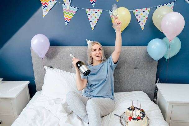 Happy emotional woman in pajama and party cap sitting on the bed with festive cake and champagne bottle in decorated bedroom. Time for yourself Celebration at home. Happy birthday concept - Φωτογραφία, εικόνα