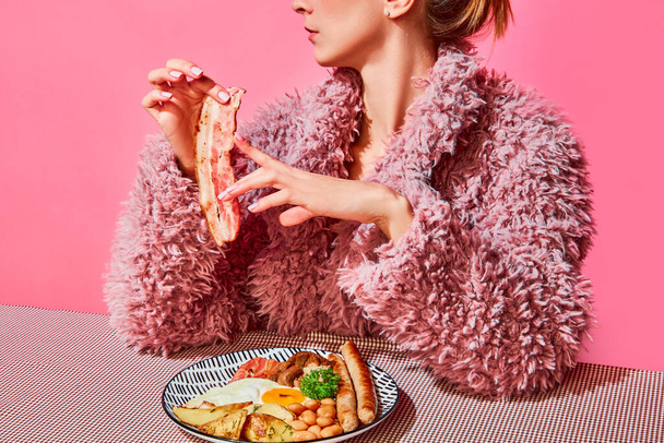 Stylish woman in pink furry coat eating american breakfast with fried eggs, sausages and bacon over pink background. Vintage, retro style. Food pop art. Complementary colors. Copy space for ad, text - Foto, immagini