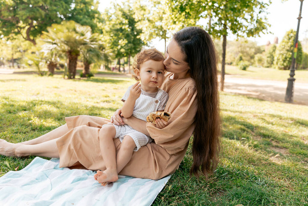 Šťastná roztomilá holčička s kudrlinkami sedící matce na kolenou a dívající se s úsměvem na kameru. Yon žena s dlouhými tmavými vlasy drží svou dceru a tráví čas v parku  - Fotografie, Obrázek