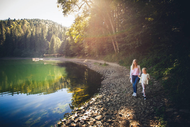 Mom and daughter walk hand in hand along the shore of a mountain lake on a bright sunny day. Coniferous forest around. - 写真・画像