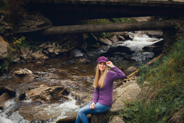 Una joven delgada con el pelo largo y rubio, con ropa de verano y un sombrero, sonríe y se sienta en la orilla de un río de montaña cerca del puente, durante el día. - Foto, Imagen