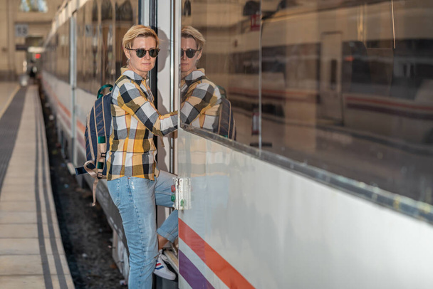 ヨーロッパで電車で旅するバックパックとサングラス付きの無地のシャツとジーンズの大人の35歳のレズビアン女性。スペインのバルセロナに駅. - 写真・画像