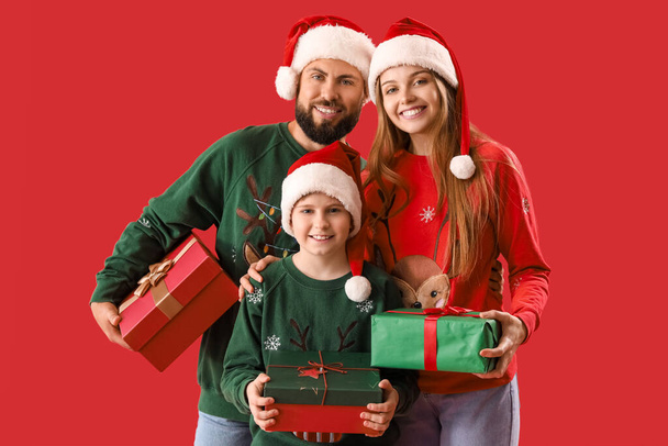 Gelukkige ouders met hun zoontje in kerstmutsen en kerstcadeaus op rode achtergrond - Foto, afbeelding