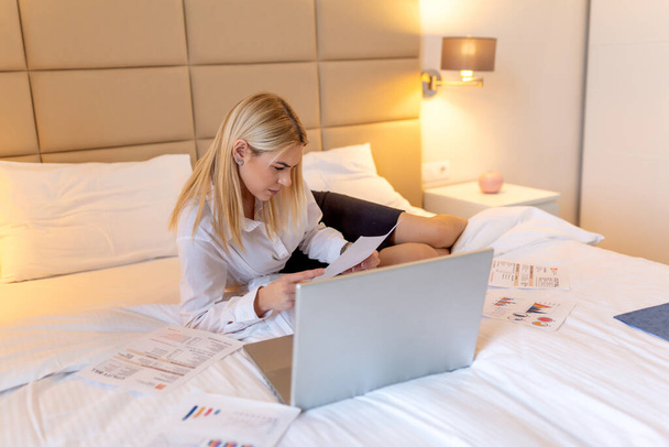 Businesswoman in suit talking on smartphone and looking at notebook in hotel room. Business Woman working from a hotel room. Beautiful young woman talking on mobile smart phone. - Φωτογραφία, εικόνα