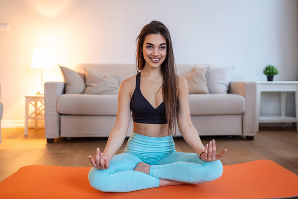 Young attractive smiling woman practicing yoga, sitting in Half Lotus exercise, Ardha Padmasana pose, working out, wearing sportswear, meditation session, indoor full length, home interior - Fotografie, Obrázek