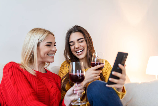 Przyjaciele z winem w domu. Szczęśliwy uśmiech młodych kobiet przyjaciół patrząc na zdjęcia w telefonie komórkowym. Dziewczyny dobrze się bawią. Dwie przyjaciółki relaks w domu z kieliszkiem wina rozmawiając razem - Zdjęcie, obraz
