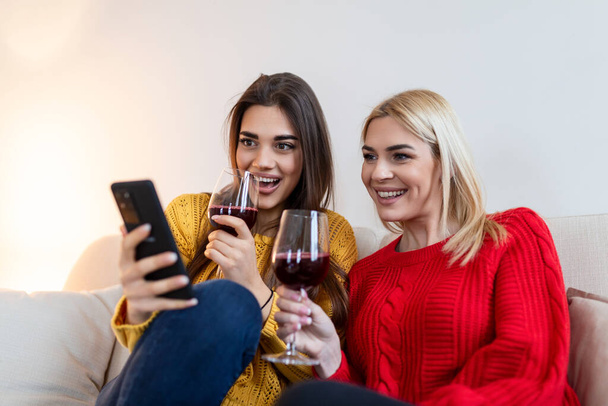 ワインと居心地の良いロフトアパートで笑うソファに座っている女性。2人の女友達が一緒にワインを飲みながら自宅でソファでリラックス. - 写真・画像