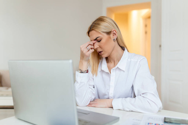 Jovem mulher frustrada trabalhando na mesa de escritório na frente do laptop que sofre de dores de cabeça diárias crônicas, tratamento on-line, apontando para uma consulta médica, radiação eletromagnética, salário doente
 - Foto, Imagem